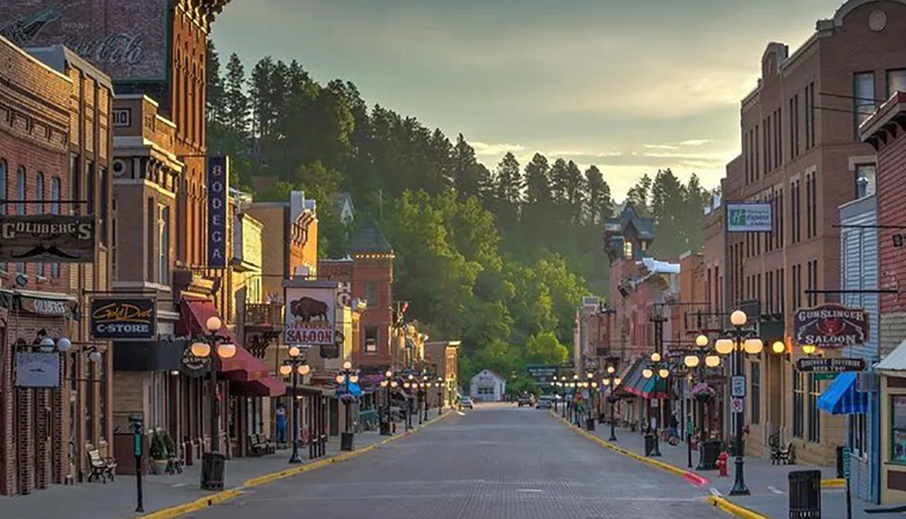 The image depicts a picturesque vintage-looking main street lined with old-fashioned storefronts and street lamps set against a backdrop of green hills and a clear early morning or late afternoon sky