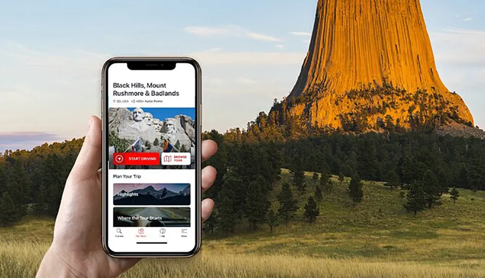 A person is holding a smartphone with a travel guide app open to a page about Black Hills Mount Rushmore  Badlands with the iconic Devils Tower visible in the background