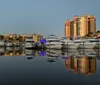 The image depicts a tranquil marina at dusk with still waters reflecting luxury boats and the surrounding upscale buildings under a fading sky