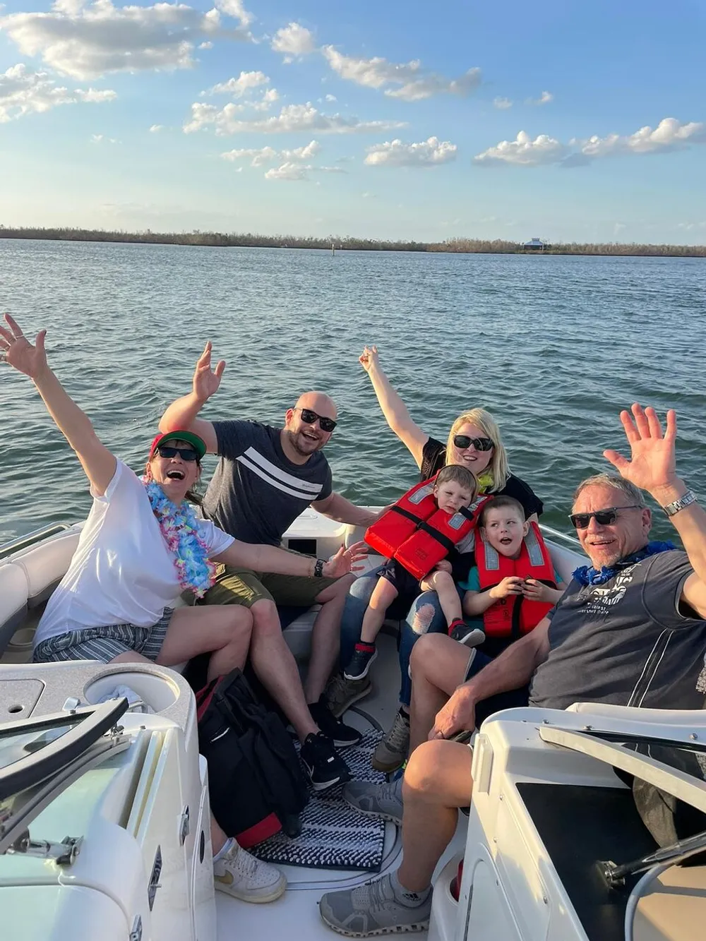 A group of six joyful people are having a good time on a boat with some of them raising their arms in the air against a backdrop of calm waters and a clear sky