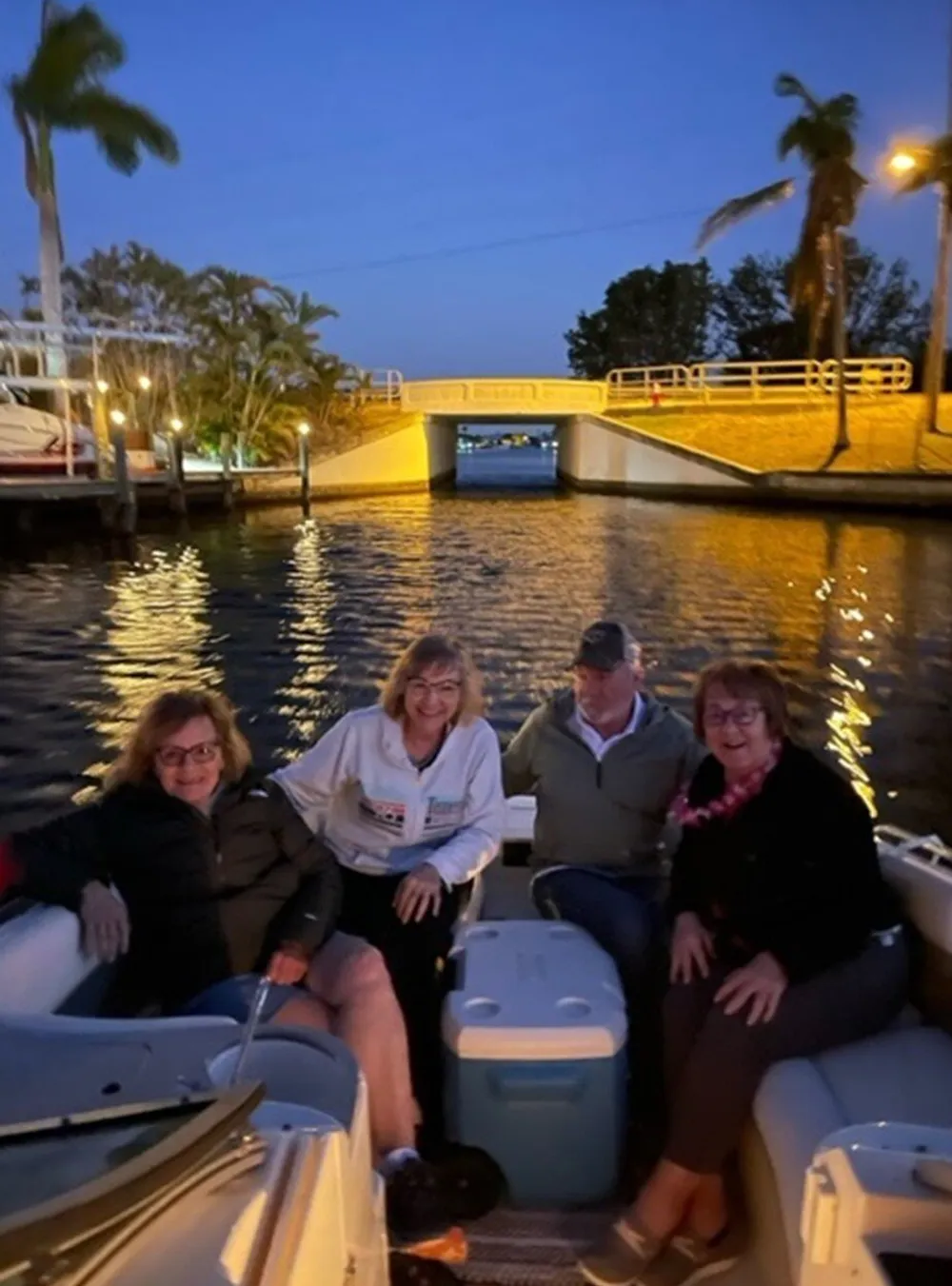 Four people are smiling for a photo while sitting on a boat in a canal with palm trees and a bridge in the background during twilight