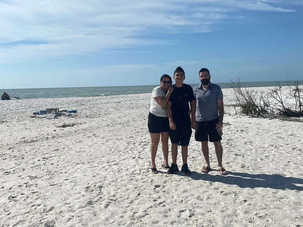 Three people are standing together smiling on a sandy beach with the ocean in the background under a clear sky