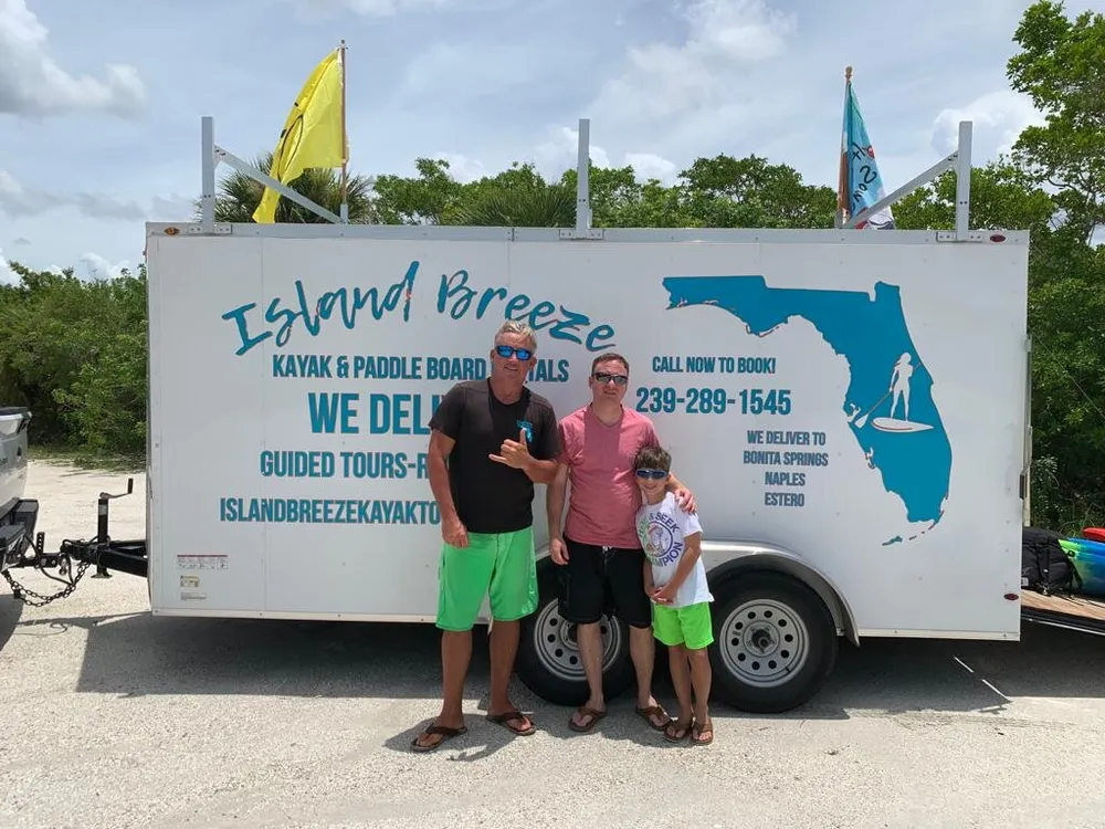 Three people are posing for a photo in front of a trailer advertising kayak and paddle board rentals and guided tours