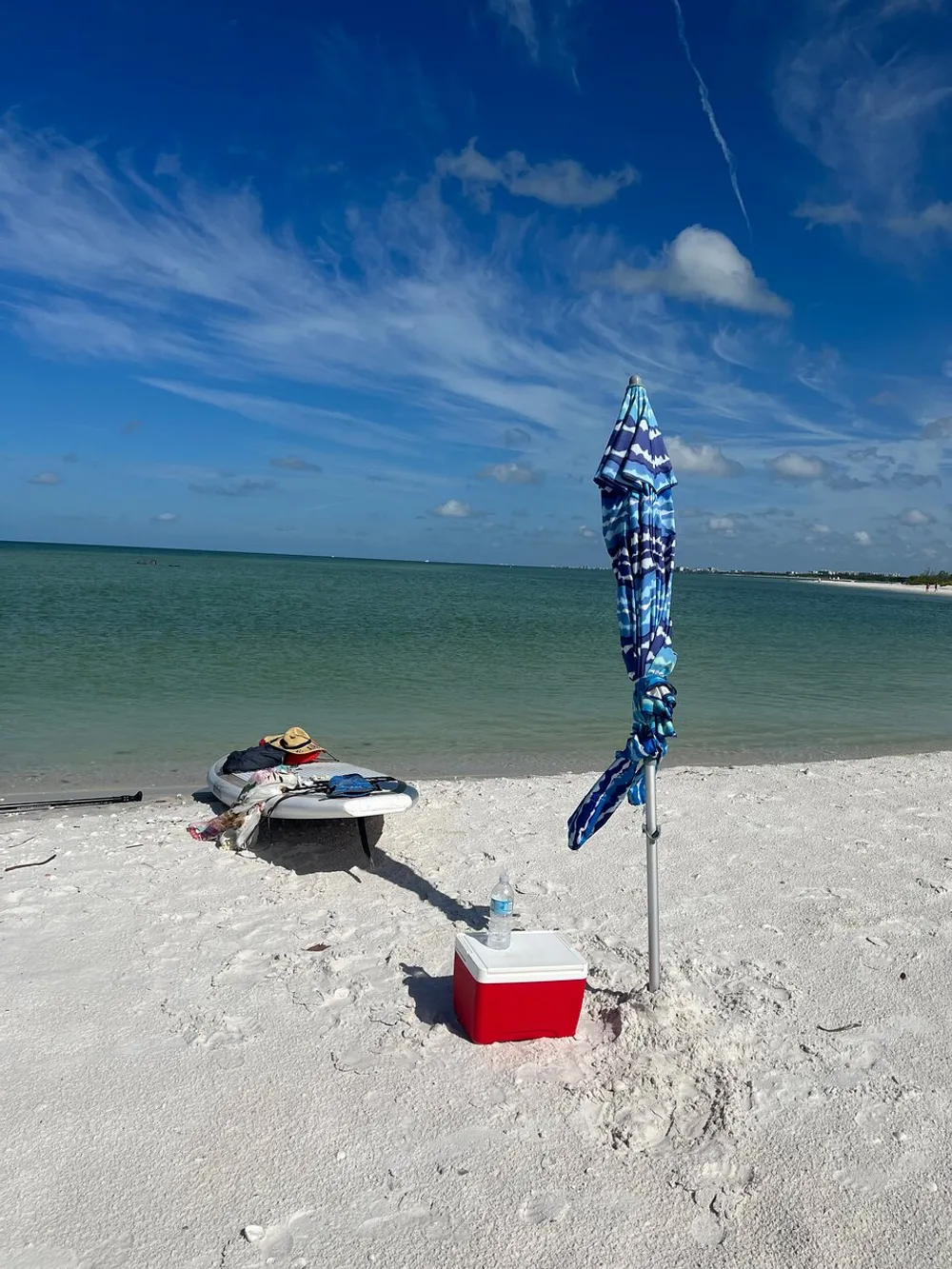 A blue and white umbrella is anchored in a sandy beach with a paddleboard hat water bottle and cooler nearby against a backdrop of calm waters and a blue sky with wispy clouds