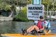 Two people in a kayak are passing by a warning sign indicating that local knowledge is required and specifying a water depth of 3.0 feet.