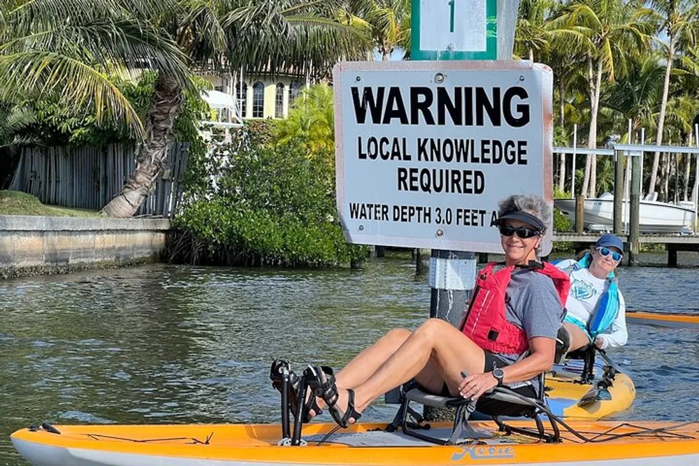 Two people in a kayak are passing by a warning sign indicating that local knowledge is required and specifying a water depth of 30 feet