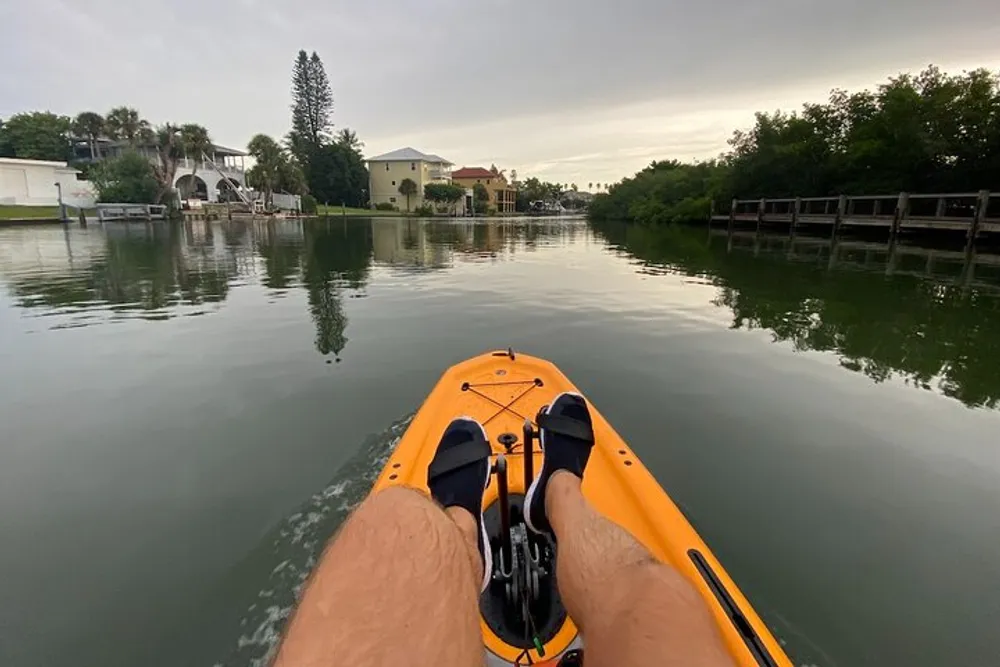 The image shows a first-person view of someone kayaking on a calm river with residential homes and docks on either side under an overcast sky