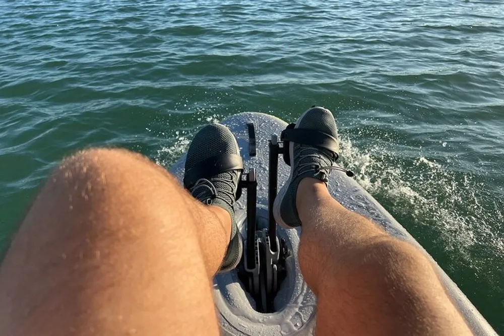 The image captures a persons point of view while seated on a watercraft with their knees and sneaker-clad feet visible against a backdrop of sparkling water