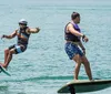 Two people are hydrofoiling on a calm sea with the person in the foreground seemingly in the process of falling off his board