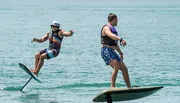 Two people are hydrofoiling on a calm sea, with the person in the foreground seemingly in the process of falling off his board.