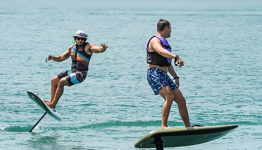 Two people are hydrofoiling on a calm sea with the person in the foreground seemingly in the process of falling off his board
