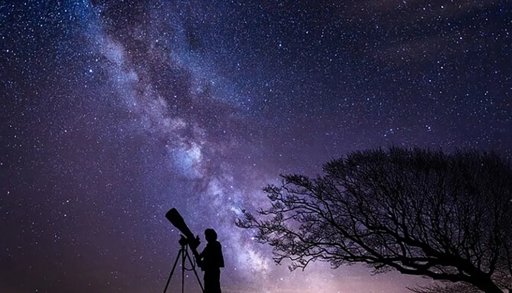 A silhouetted person observes the night sky filled with stars and the Milky Way through a telescope
