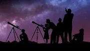 A group of silhouetted people, including children and adults, are stargazing with telescopes under a starry night sky.
