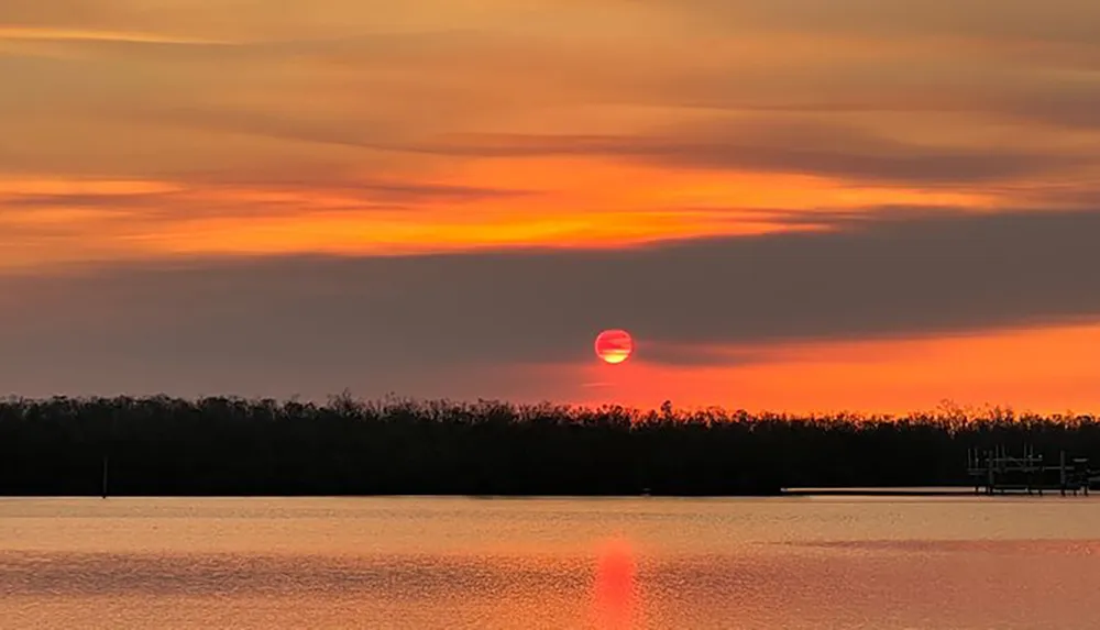 The image captures a serene sunset with a vivid orange sun partially obscured by cloud layers reflecting on a calm water body against a silhouette of trees