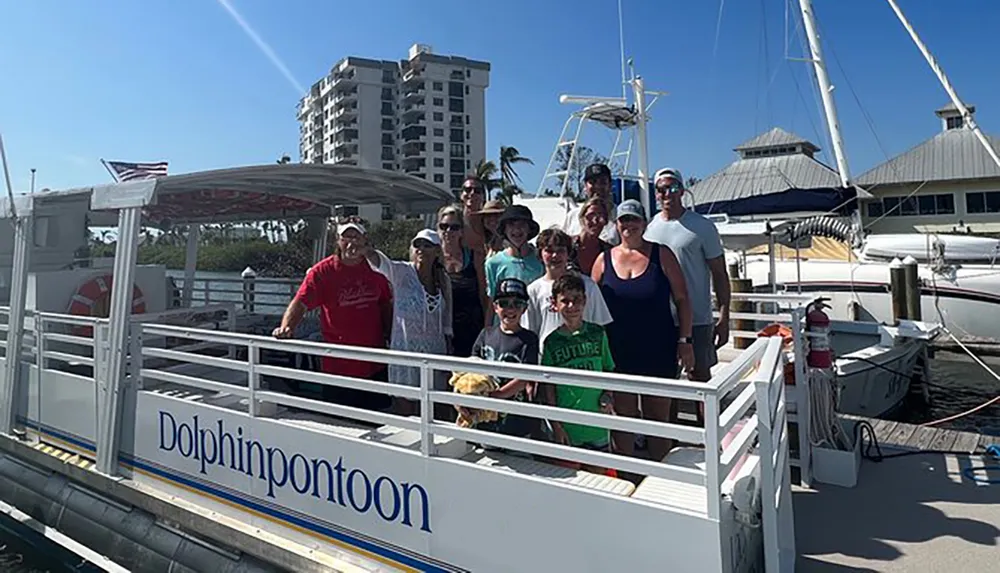 A group of people is posing for a photo on a sunny day on the deck of a boat named Dolphin Pontoon docked at a marina