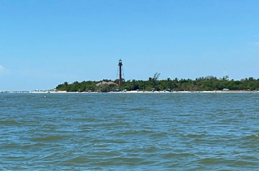 The image shows a coastal scene with a tall lighthouse standing near the waters edge viewed from across a body of water with gentle waves under a clear blue sky