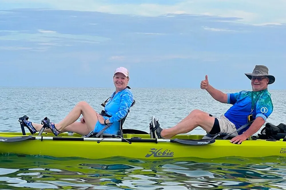 Two people are smiling and enjoying themselves on a tandem kayak in calm waters with the person at the back giving a thumbs up