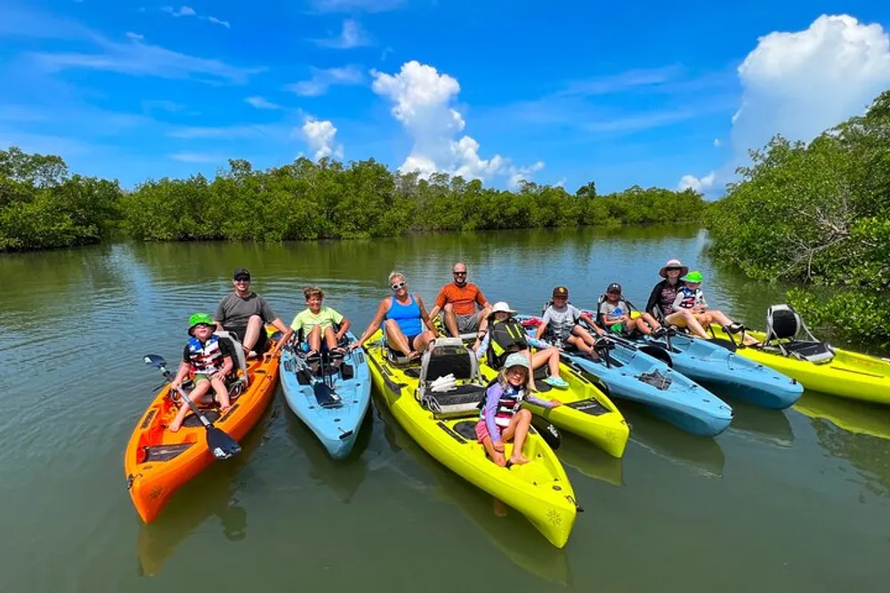 A group of people including adults and children are smiling and posing for a photo while sitting in colorful kayaks on a calm river surrounded by greenery