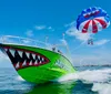 A brightly colored green boat with a shark face painted on it is towing a parasail with two people flying above the water against a clear blue sky