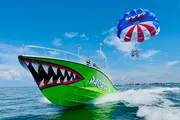 A brightly colored green boat with a shark face painted on it is towing a parasail with two people flying above the water against a clear blue sky.