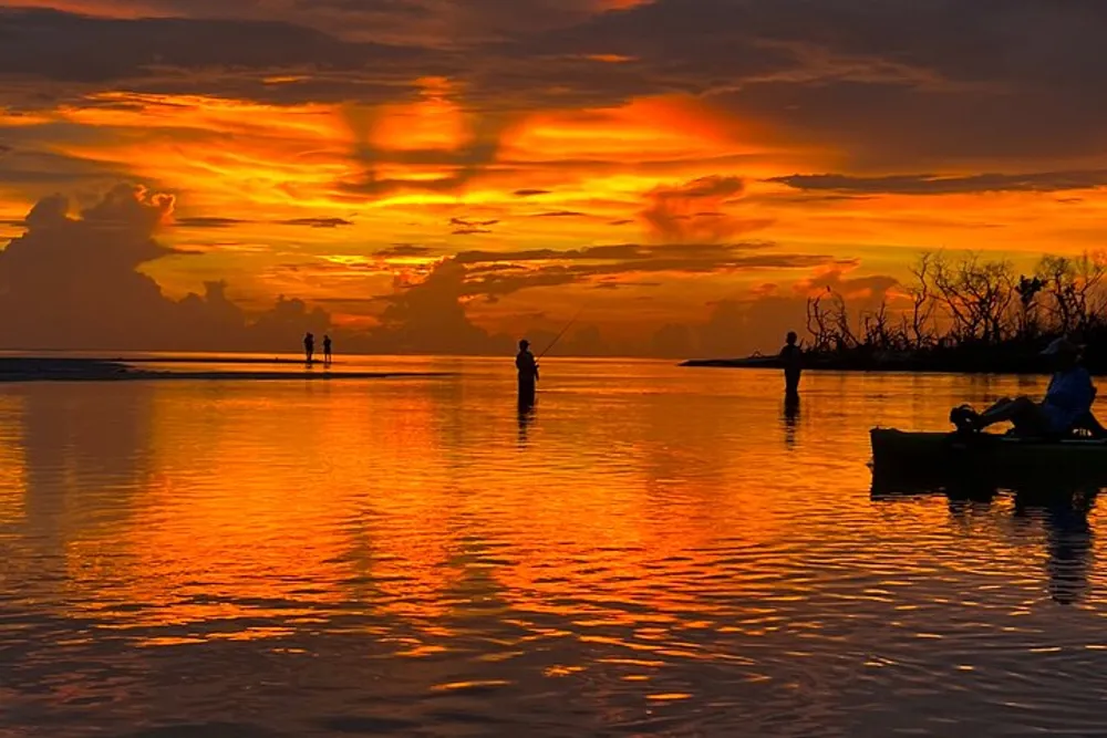 A vibrant sunset casts a fiery glow over the water where people are silhouetted against the sky while fishing and paddling a canoe
