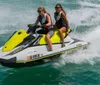 Two people are riding a yellow and white jet ski on a sunny day with clear water splashing around
