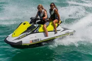 Two people are riding a yellow and white jet ski on a sunny day with clear water splashing around.