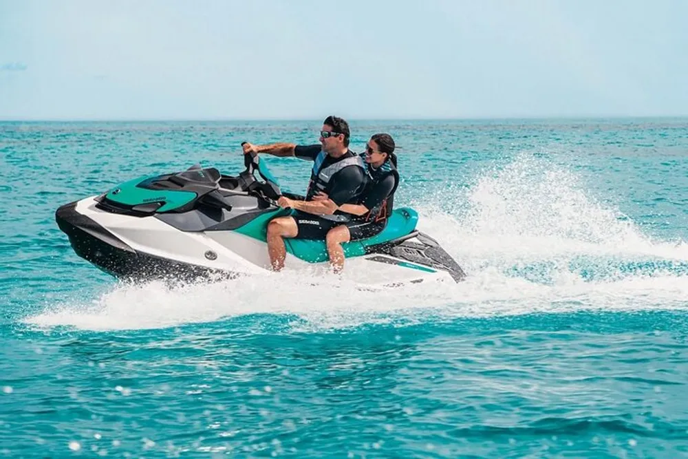 Two people are riding a jet ski across a clear blue body of water creating a spray of water in their wake