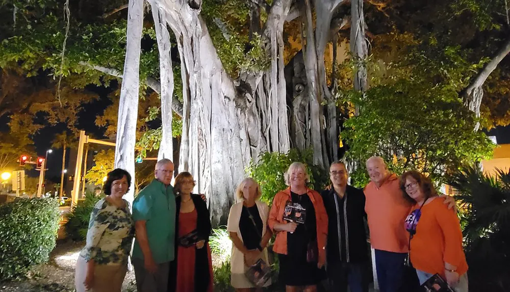 A group of people is posing for a picture at night in front of a large Banyan tree with intricate roots and branches