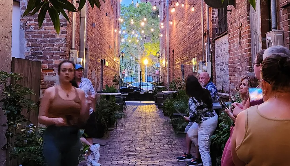 The image captures a lively evening in a quaint alleyway adorned with string lights where people are gathered and engaging in conversation