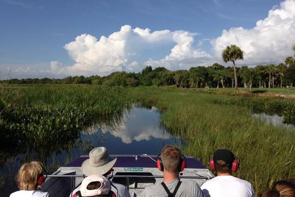 Passengers are enjoying a scenic airboat tour through a lush green wetland under a blue sky dotted with white clouds