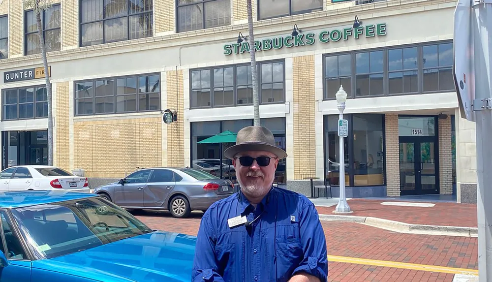 A smiling person in a fedora hat and sunglasses is standing in front of a Starbucks Coffee shop on a sunny day