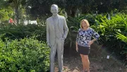 A person is standing next to a statue of a man in a garden setting, smiling at the camera.