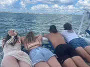 Four friends are lying on their stomachs on a net of a catamaran, enjoying the ocean view with a coastline in the background.