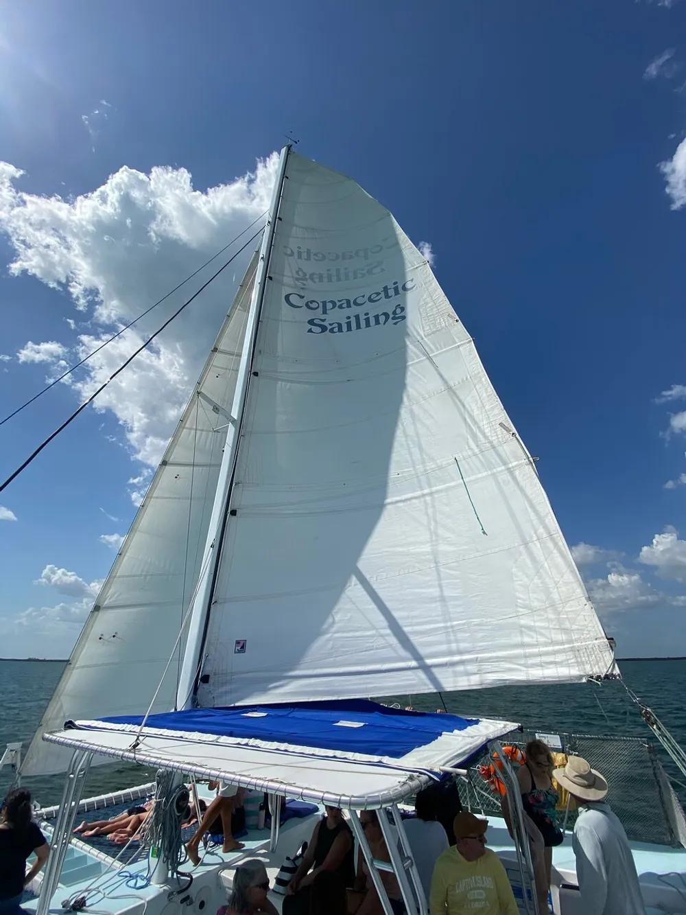 A group of people enjoy a sunny day on a sailboat with a large white sail set against a clear blue sky with some clouds