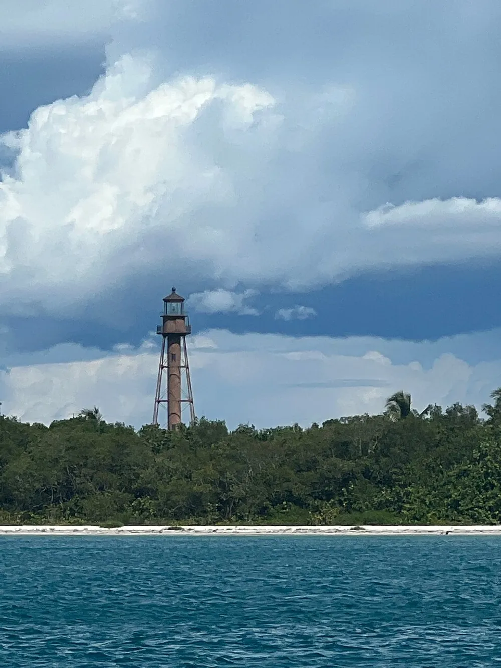 The image shows a tall slender lighthouse on a lush coastline against a backdrop of dynamic blue skies and puffy white clouds as viewed from the water