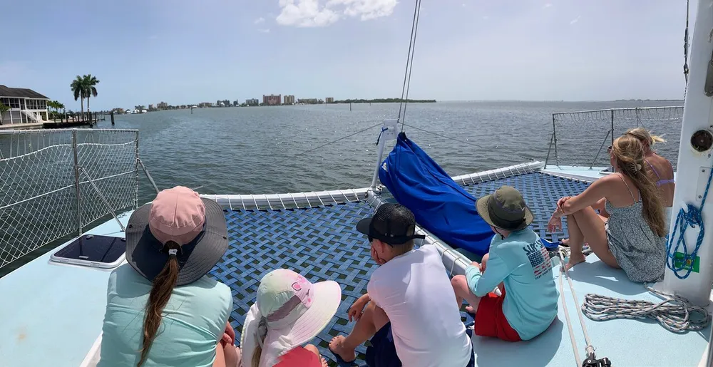 A group of people is enjoying a sunny day on the netted deck of a catamaran with calm waters and coastal buildings in the background