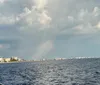 The image shows a coastal city skyline under a cloudy sky with a faint rainbow touching down on the water