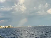 The image shows a coastal city skyline under a cloudy sky with a faint rainbow touching down on the water.