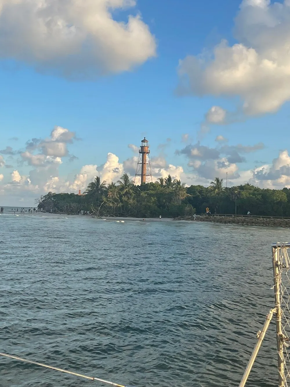The image captures a rustic lighthouse standing among tropical foliage on a coastal area with the ocean in the foreground and a partly cloudy sky above