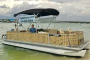 A person is sitting on a pontoon boat adorned with a tiki bar design, docked in calm waters with a bridge in the background.