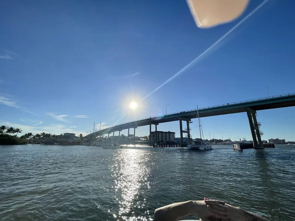 The image depicts a sunlit scene with a bridge crossing over a body of water boats moored nearby and a clear blue sky with the sun creating a starburst effect