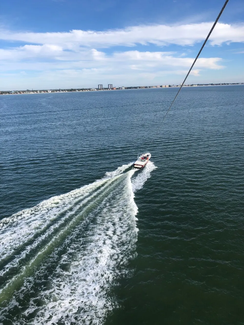 A boat is cruising through the water leaving a foamy wake with a coastline and buildings in the background and a rope diagonally entering the frame from the top