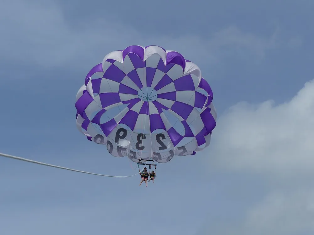 A purple and white parasail with two people suspended underneath soars against a blue sky with scattered clouds