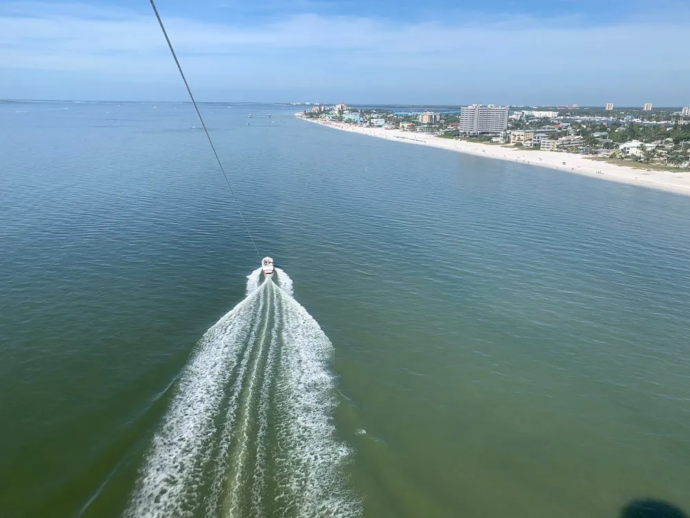 The image depicts a scenic view of a coastline with a boat creating a wake in the water likely taken from the perspective of someone parasailing above the sea