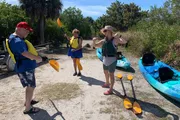 Three people appear excited and ready for a kayaking adventure, standing near blue kayaks and holding paddles outdoors on a sunny day.