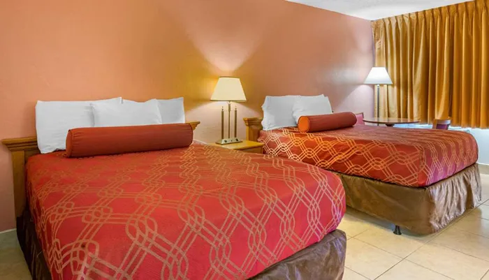 The image shows a standard hotel room with two double beds featuring red patterned bedspreads white pillows a bedside table with a lamp and golden curtains against an orange wall