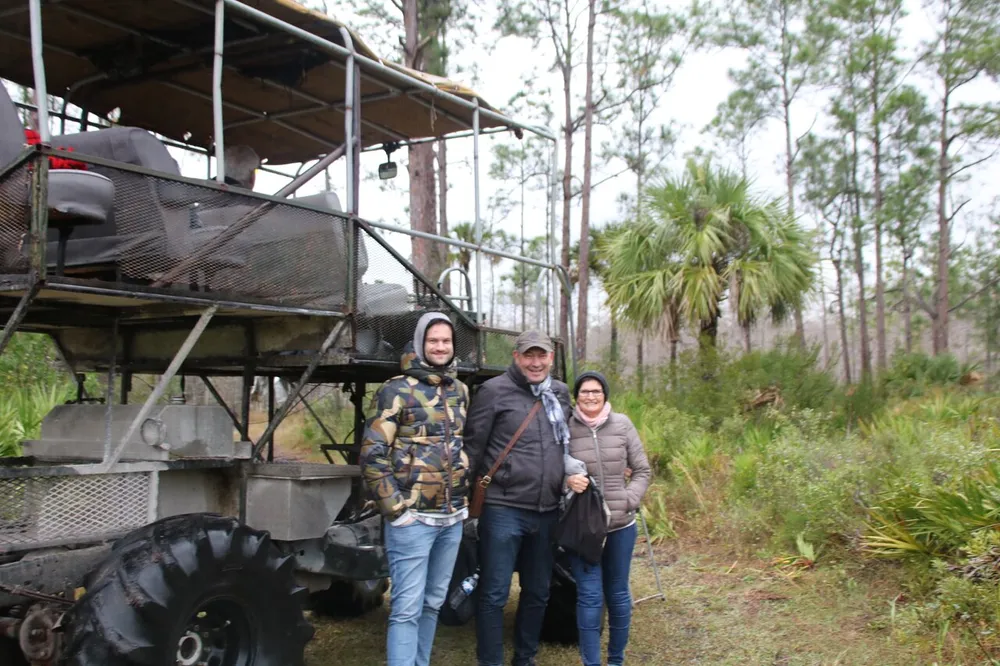 Three people are posing with smiles in front of a large all-terrain vehicle in a forested area