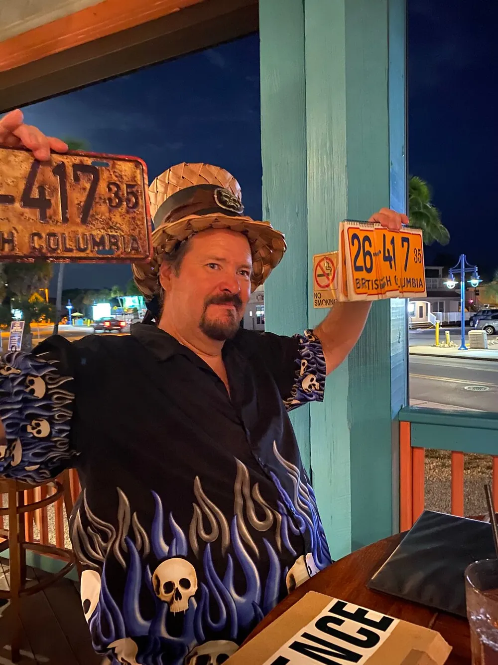 A person wearing a puffy pirate hat and a flame-pattern shirt with skull motifs is holding up two weathered license plates from British Columbia