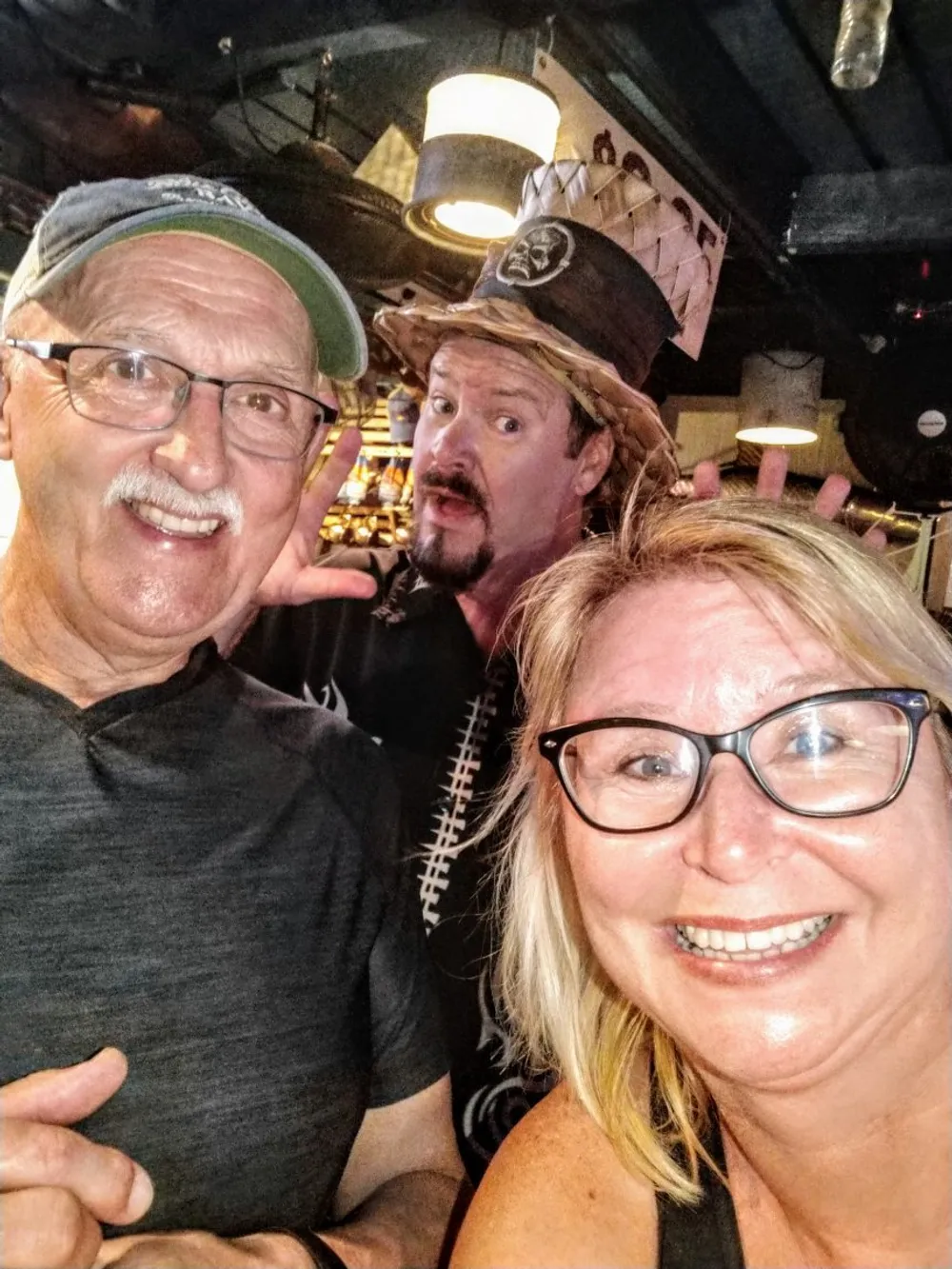 Three people are posing for a cheerful selfie with one person wearing a pirate hat and striking a playful pose in the background
